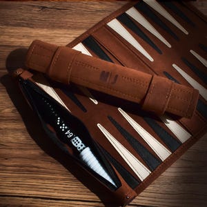 Könnte beinhalten: Ein braunes Backgammon-Set mit Reißverschlusstasche, Würfeln und einem Würfelbecher. Das Set enthält eine braune Ledertasche mit schwarzen und weißen Dreiecken. Der Würfelbecher ist weiß und schwarz, und das Etui hat die Initialen "MJ".