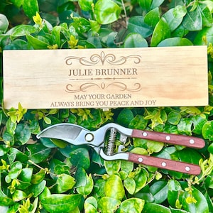 May include: A wooden box with the name "JULIE BRUNNER" and the text "MAY YOUR GARDEN ALWAYS BRING YOU PEACE AND JOY" is displayed with a pair of gardening shears. The shears have a silver blade and brown wooden handles.