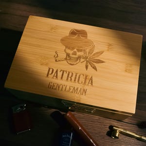 Peut inclure: Un coffret en bambou avec un crâne portant un fedora, fumant une cigarette et une feuille de cannabis. La boîte est gravée du nom "PATRICIA" et du mot "GENTLEMAN". Un briquet, un cigare et une pipe sont à côté de la boîte.