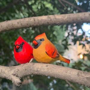 May include: Two wooden cardinal bird figurines, one bright red and the other orange, perched on a tree branch. The birds have black beaks and eyes, with detailed carving. The background is blurred green foliage.
