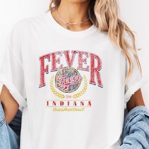 May include: White t-shirt with a distressed graphic featuring the word "FEVER" in red, arched above a circular emblem and the words "INDIANA Basketball" in yellow. The shirt has rolled-up sleeves and is paired with a denim jacket.