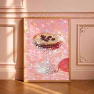 Könnte beinhalten: Gerahmter Druck mit einem Kaffee-Cocktail in einem Glas im Discokugel-Stil. Das Getränk hat eine schaumige Oberfläche mit Kaffeebohnen. Der Hintergrund ist rosa mit glitzernden Glitzer. Der Rahmen ist goldfarben, und die Wand ist rosa.