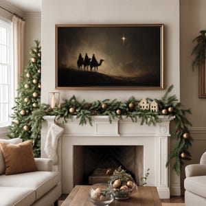 May include: A framed artwork depicting three figures on camels, illuminated by a star, hangs above a fireplace decorated with a garland, ornaments, and miniature houses. A Christmas tree with gold ornaments stands to the left, creating a festive holiday scene.