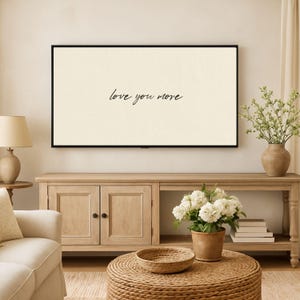 May include: A framed wall art piece with the phrase "love you more" in elegant script. The artwork is displayed above a wooden cabinet, complemented by a round woven coffee table, a vase of flowers, and a lamp.