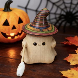 May include: A wooden ghost figurine wearing a colorful witch hat and holding a small broom. A carved pumpkin with a lit interior and fall leaves are in the background. A black spider and web are also in the background. The figurine has a smiling face.
