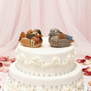 Könnte beinhalten: Eine zweistöckige weiße Hochzeitstorte, verziert mit weißen Zuckergussrosen und silbernen Perlen. Zwei geschnitzte Holzfiguren von Enten, eine braune und eine graue, sitzen auf der Torte. Die Enten blicken sich an.