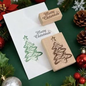 Puede incluir: Imagen navideña con una tarjeta blanca con un sello de árbol de Navidad verde y las palabras "Merry Christmas". También hay dos sellos de madera con el mismo texto y un diseño de árbol de Navidad. Decoraciones festivas.