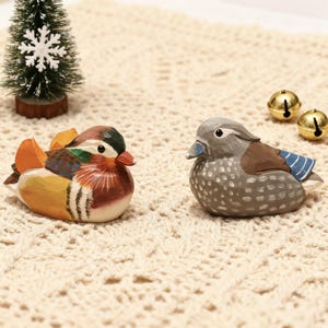 Könnte beinhalten: Zwei handgeschnitzte Holzfiguren von Enten. Eine ist farbenfroh mit orangefarbenen, gelben, roten und grünen Akzenten. Die andere ist grau, braun und blau. Ein kleiner Weihnachtsbaum und zwei goldene Glocken sind im Hintergrund.