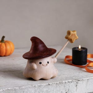 Pode incluir: Estatueta de madeira de um fantasma sorridente usando um chapéu de bruxa marrom escuro e segurando uma varinha em forma de estrela. Uma pequena abóbora laranja e uma vela preta com uma fita laranja estão no fundo. Decoração de Halloween.