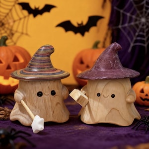 May include: Two wooden Halloween-themed figurines, each with a witch hat. One hat is striped, the other is purple. The figurines have smiling faces and hold a broom and a block. Pumpkins and bats decorate the background.