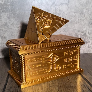 Puede incluir: Una caja rectangular dorada con una tapa en forma de pirámide. La pirámide y la caja están adornadas con diseños y símbolos intrincados. La pirámide presenta un prominente símbolo de ojo. La caja es probablemente un artículo de colección o decorativo.