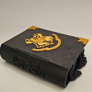 May include: A black Harry Potter book-shaped box with gold detailing. The box has the Harry Potter logo and text on the spine. The top features a gold emblem and corner details. Likely a decorative storage item.
