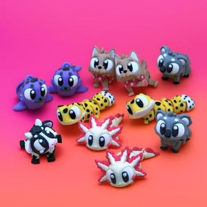 Peut inclure: Plusieurs figurines miniatures d'animaux colorés, dont des chats, des koalas et des axolotls, sont disposées sur un fond dégradé rose et orange. Les figurines sont détaillées avec divers motifs et couleurs, créant une esthétique ludique et fantaisiste.