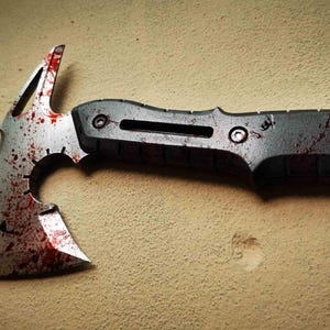 Könnte beinhalten: Eine silberne und schwarze taktische Tomahawk-Axt mit blutbespritzter Klinge. Die Axt hat eine scharfe Klinge, einen Hammerkopf und einen strukturierten Griff. Das Design deutet auf ein Überlebens- oder taktisches Werkzeug hin.