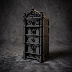 Puede incluir: Un pequeño mueble negro de estilo gótico. Cuenta con cinco cajones con tallas decorativas y tiradores con cabeza de león. La parte superior tiene un diseño arqueado. El mueble se encuentra sobre un fondo gris oscuro, creando un efecto dramático.