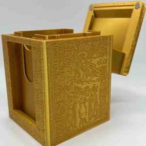 Puede incluir: Una caja rectangular dorada con tapa abierta. La caja está adornada con obras de arte grabadas detalladas que representan figuras y jeroglíficos. La tapa está unida con una bisagra y tiene un cierre magnético. La caja está diseñada para guardar objetos.