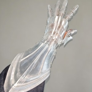 May include: A clear, articulated hand-shaped sculpture. The sculpture is made of transparent plastic with silver-colored rivets. The fingers are segmented and pointed, and the wrist area is flared. The hand is posed in a gesture.