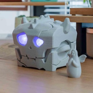 May include: A grey, skull-shaped decorative box with glowing blue eyes. The box has a hinged design and a textured surface. A small, white angel figurine stands beside the box. A unique home decor item.