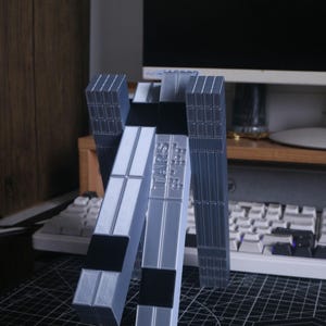 May include: A 3D-printed stand in shades of gray and black, featuring the text "TARS RS". The stand has a geometric design with rectangular and square elements. It is designed to hold a device, possibly a phone or tablet.