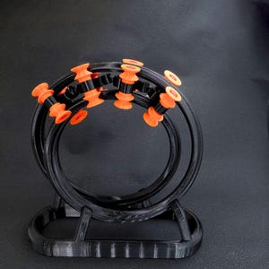 May include: A black, 3D-printed sculpture featuring two intersecting circular forms. The sculpture has orange and black accents, with several orange cylindrical pieces evenly spaced around the black rings. The base is also black.