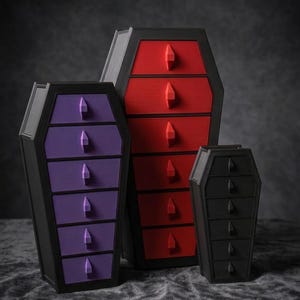 Puede incluir: Tres arcones de almacenamiento con forma de ataúd de diferentes tamaños. Los arcones son negros con cajones rojos, morados y negros. Cada cajón tiene un tirador en forma de gema. Los arcones están colocados sobre una superficie oscura y texturizada.