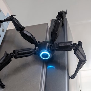 Può includere: Un ragno robotico nero stampato in 3D con zampe articolate e un corpo centrale. Il corpo presenta un anello blu luminoso ed è montato su un frigorifero. Le zampe del ragno sono posizionate per afferrare.