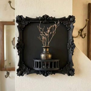 Peut inclure: Étagère murale noire ornée de forme carrée. Elle comprend une petite étagère décorative supportant un vase avec des fleurs séchées. Le vase a une finition dorée métallique. L'étagère est fixée sur un mur crème.