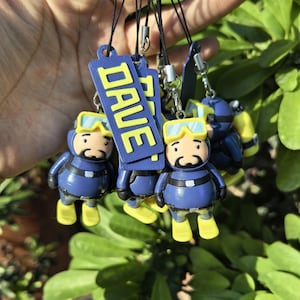Peut inclure: Plusieurs porte-clés sur le thème de la plongée, bleus et jaunes. Chaque porte-clés représente une figurine de plongeur avec un masque et des palmes jaunes, et une étiquette bleue avec le mot "DAVE". Les porte-clés sont attachés à des lanières noires.