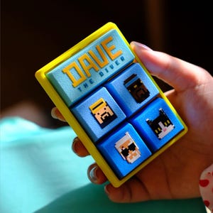 Peut inclure: Une œuvre d'art pixélisée encadrée en jaune avec le texte "DAVE THE DIVER" en orange. La pièce présente quatre carrés bleus avec des personnages en pixel art. Les personnages incluent un plongeur, un chat et deux autres personnages.