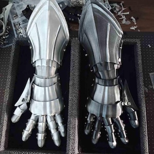 May include: Two metallic silver gauntlets with articulated fingers, designed with layered plates and a pointed top. One gauntlet is fully silver, the other is a gradient of silver and black. The gauntlets are displayed on a dark surface.