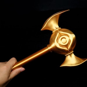 Könnte beinhalten: Ein goldfarbener, verzierter Stab oder Zepter mit einem zentralen kugelartigen Design und zwei gebogenen, flügelartigen Vorsprüngen. Der Stab wird vor einem schwarzen Hintergrund gehalten, der seinen metallischen Glanz und seine komplizierten Details hervorhebt. Das Objekt scheint ein Requisit oder ein dekorativer Gegenstand zu sein.