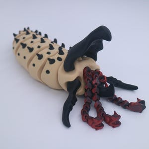 Op de afbeelding: Een beige, gesegmenteerd beeldhouwwerk van een wezen met zwarte stekels, zwarte aanhangsels en rode, kettingachtige elementen. Het beeldhouwwerk heeft een gedetailleerd, gestructureerd uiterlijk tegen een witte achtergrond.