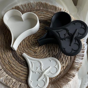 Peut inclure: Trois plateaux décoratifs en forme de cœur, blancs et noirs, disposés sur un dessous de plat circulaire tissé. Un plateau blanc a une forme de cœur simple, tandis que les deux autres présentent des motifs gothiques ornés. Les plateaux sont en matériau solide.