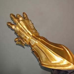 May include: A golden, articulated gauntlet, likely for cosplay or display. The metallic hand armor features individual finger segments and overlapping plates, riveted together. The gauntlet is a bright, polished gold color, reflecting light.