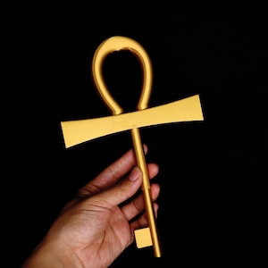 Könnte beinhalten: Ein goldfarbenes Ankh-Symbol, eine kreuzartige Form mit einer Schlaufe oben, wird vor einem schwarzen Hintergrund gehalten. Das Ankh ist ein Symbol für Leben und Unsterblichkeit im alten Ägypten.
