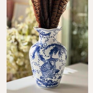 May include: A white and blue ceramic vase with a monkey design, filled with brown feathers. The vase has a ruffled top and a detailed blue pattern. The vase is on a white surface.