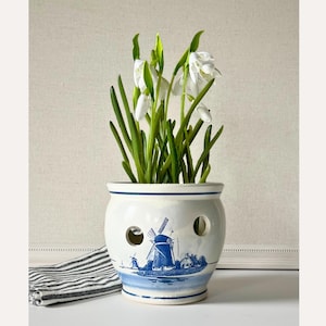 May include: A white ceramic planter with a blue windmill design, holding white flowers with green stems. The planter has two circular holes and a blue rim. A striped cloth is on the left.
