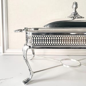May include: A silver-colored chafing dish with a lid and ornate detailing. The dish has a rectangular shape with decorative openwork sides and stands on curved legs. The lid features a decorative handle. The chafing dish is designed for serving food.
