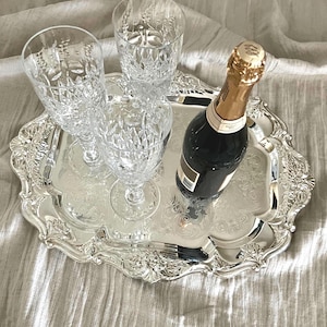 May include: A rectangular silver-toned serving tray with ornate embossed edges and a central floral pattern. The tray holds two clear, textured crystal glasses and a dark glass bottle with a gold foil-wrapped neck and a tan label.