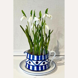 Op de afbeelding: Een blauw-wit patroon kruik met een handvat, gevuld met sneeuwklokjes. De kruik staat op een bijpassende schotel. De sneeuwklokjes hebben witte bloemblaadjes en groene stelen en bladeren. De kruik heeft een blauw-wit geometrisch ontwerp.