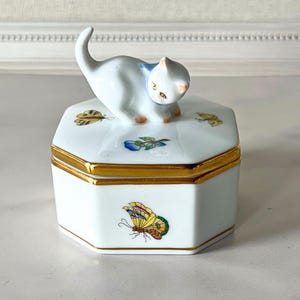 May include: A white porcelain trinket box with a gold rim and a cat figurine on top. The box is decorated with colorful butterfly and floral designs. The cat has orange paws and a blue collar.