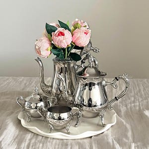 May include: A silver-plated tea set consisting of a teapot, a lidded sugar bowl, and a creamer, all featuring ornate handles and feet. A bouquet of pink artificial peonies with green leaves is arranged inside the tall, slender teapot.