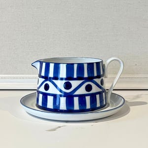 Op de afbeelding: Een blauw-witte keramische creamer en schotel set. De creamer heeft een handvat en tuit, met een ontwerp van verticale blauwe strepen en een golvend patroon met blauwe stippen. De schotel is wit met een blauwe rand.