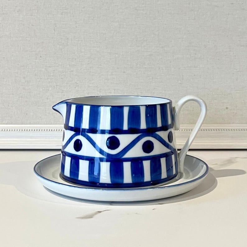 Op de afbeelding: Een blauw-witte keramische creamer en schotel set. De creamer heeft een handvat en tuit, met een ontwerp van verticale blauwe strepen en een golvend patroon met blauwe stippen. De schotel is wit met een blauwe rand.
