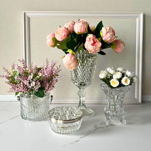 May include: A collection of clear glass vessels including a tall diamond-cut pedestal vase with pink peonies, a ribbed cylindrical vase with lavender, a small footed vase with white flowers, and a round lidded glass jar with a silver rim.