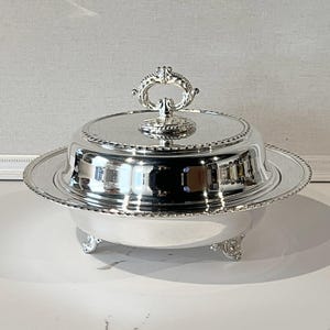 May include: A silver-plated serving dish with a domed lid and ornate handle. The dish has a round base with decorative feet and a beaded edge. The lid features a detailed, scroll-like handle. The reflective surface of the dish creates a mirror-like effect.