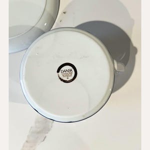 Op de afbeelding: Witte keramische kom met een blauwe rand en een handvat. De onderkant van de kom heeft een zwart en bruin cirkelvormig logo met de tekst "DANSK INTERNATIONAL DESIGNS LTD PHILIPPINES".