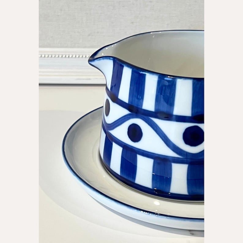 Op de afbeelding: Een wit en blauw keramisch melkkan- en schoteltje. De melkkan heeft verticale blauwe strepen en een decoratief blauw patroon met stippen. Het schoteltje heeft een blauwe rand. De set is bedoeld voor het serveren van room of melk.