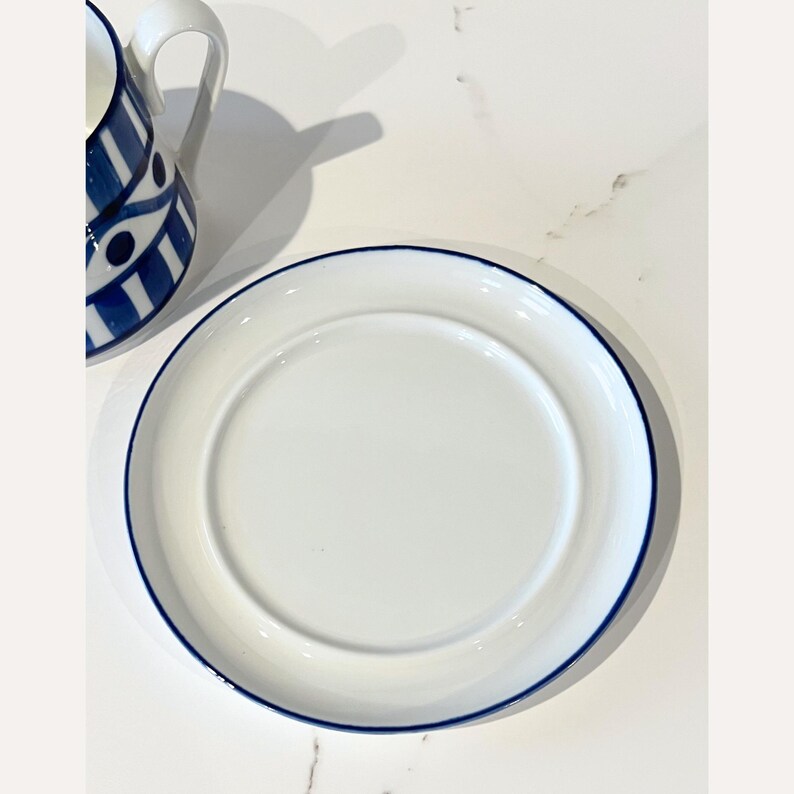 Op de afbeelding: Witte keramische plaat met een blauwe rand. Een witte keramische mok met een blauw en wit geometrisch patroon is zichtbaar. De plaat is rond en heeft een licht verhoogde rand.