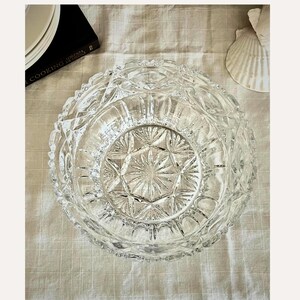 Peut inclure: Un bol en verre taill&eacute; transparent avec un motif en &eacute;toile au centre et un motif taill&eacute; en diamant sur le bord. Le bol repose sur une surface textur&eacute;e de couleur claire. Un livre de cuisine et un coquillage sont partiellement visibles en arri&egrave;re-plan.